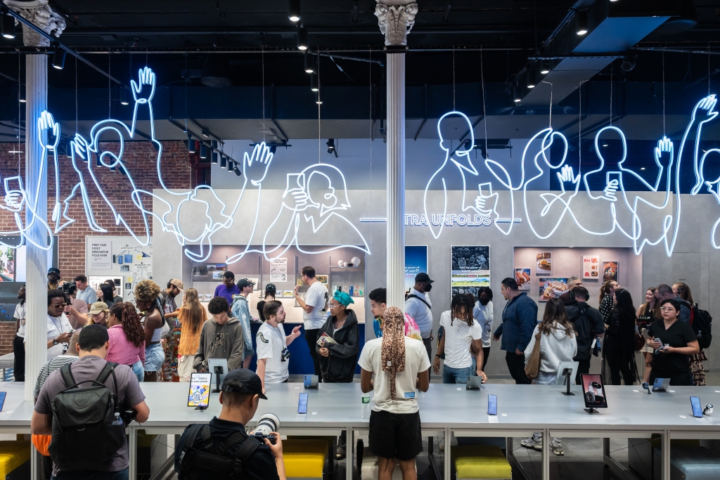 Vista interior de un espacio de experiencia de Samsung abarrotado de gente durante el lanzamiento global del Galaxy Z Fold7, Z Flip7 y la serie Galaxy Watch8, con letreros de neón artísticos en el techo que representan siluetas humanas usando teléfonos inteligentes.